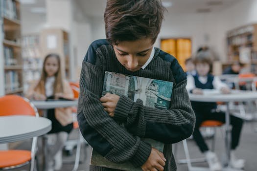 Boy stands alone, hugging book in a classroom, expressing sadness and isolation.